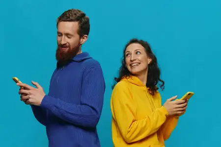 Весёлая пара — женщина и мужчина — с телефонами в руках, у них кривые улыбки, они радостны на голубом фоне
