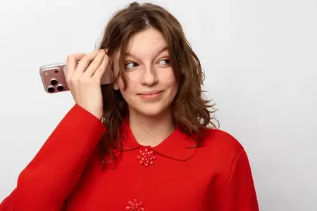 Улыбающаяся девушка с вьющимися волосами в красном свитере слушает смартфон, проявляя интерес и вовлеченность