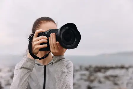Молодая женщина-фотограф в серой куртке, глубоко поглощенная съемкой прибрежного пейзажа, демонстрирует сосредоточенность, творчество и природную красоту у моря