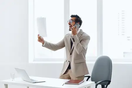 Счастливый мужчина в деловом костюме общается по телефону, сидя за ноутбуком в современном офисе