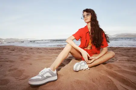 Улыбающаяся женщина в модном наряде сидит на песке на пляже, наслаждаясь беззаботным отпуском: пляжное счастье