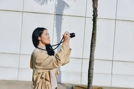 Сцена уличной фотографии: улыбающаяся женщина с фотоаппаратом, запечатлевающая яркий дневной момент рядом с деревом, повседневная одежда, образ жизни и вдохновение для путешествий