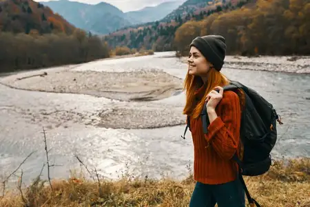 Женщина в красном свитере и кепке с рюкзаком за спиной в горах у реки на природе