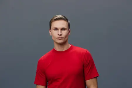Профессиональный студийный портрет уверенного молодого человека в красной футболке, смотрящего прямо в камеру со спокойным выражением лица на однотонном сером фоне