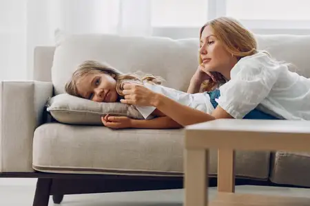 Мать и дочь общаются на уютном диване на фоне минималистичной белой стены, вместе смеются и отдыхают