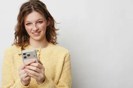 Счастливая женщина в желтом вязаном свитере держит смартфон обеими руками, мягко улыбаясь в камеру