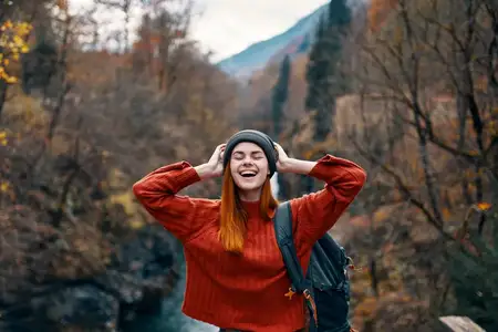 На улице стоит жизнерадостная женщина с рюкзаком на фоне гор и реки