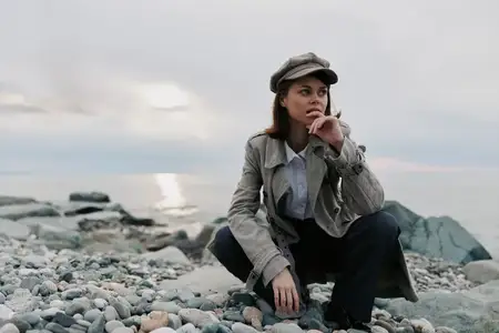 Модная молодая женщина в пальто и шляпе сидит на гальке у моря под облачным небом