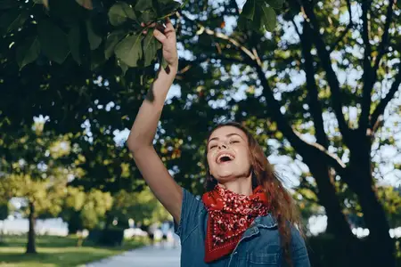 Радостная женщина, ведущая активный образ жизни, одетая в современную джинсовую уличную одежду в стиле бохо-вестерн с красным шарфом, наслаждается моментом на улице под зелеными деревьями