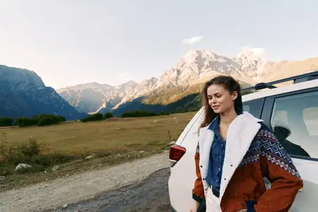 Женщина опирается на машину на обочине дороги с горным пейзажем для приключенческого путешествия, природный пейзаж и открытая дорога