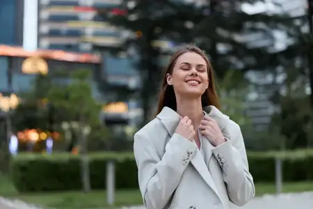 Молодая женщина в светлом пальто улыбается с закрытыми глазами, выражая расслабленные эмоции на открытом воздухе в городском парке, демонстрируя осенний стиль, современный образ жизни и позитивное…