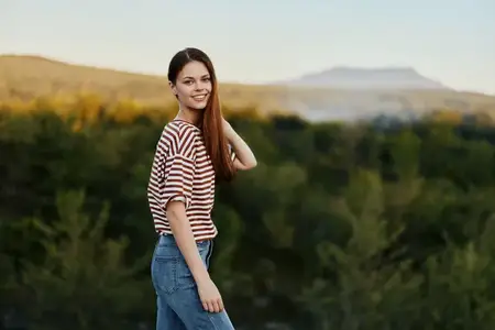 Молодая женщина смеется и смотрит в камеру, одетая в простую одежду, на фоне красивого осеннего пейзажа с горами и деревьями
