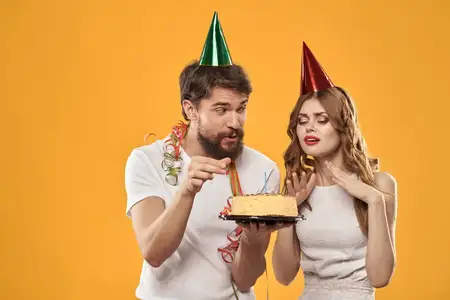 Счастливые мужчина и женщина в колпаках празднуют день рождения на жёлтом фоне, держа в руках торт