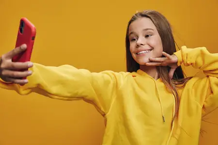 Улыбающаяся юная девушка в яркой кофте держит телефон для селфи, делая жест на желтом фоне со счастливым выражением лица для личного фото или аватара в интернете