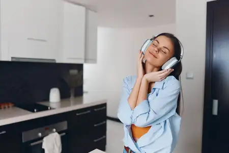 Женщина наслаждается музыкой на кухне, стоя с руками на голове