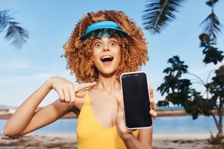 Радостная молодая женщина с вьющимися волосами держит смартфон на пляже