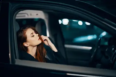 Стильная, роскошная женщина сидит в черном автомобиле ночью, поправляя свои длинные уложенные волосы и закрывая глаза от удовольствия