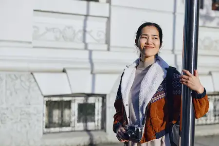 Сцена уличной фотографии: улыбающаяся женщина держит камеру рядом с каменным фасадом, модная верхняя одежда, городская среда, непринужденное и художественное повествовательное настроение сегодня