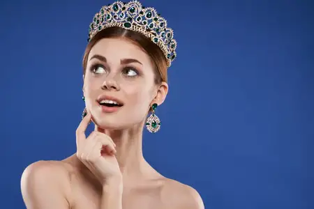 Красивая женщина в короне на голове, модель с макияжем, делает жест рукой на синем фоне