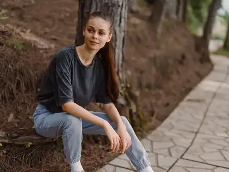 Стильная молодая женщина с длинными волосами, одетая в удобную футболку и джинсы, улыбается, сидя у дорожки в спокойной природной обстановке