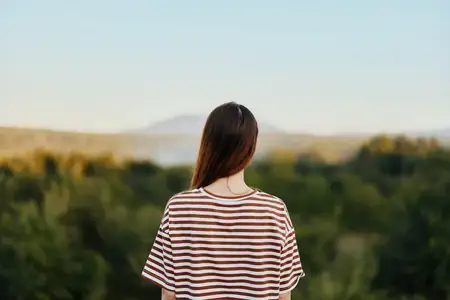 Женщина на природе стоит спиной к камере возле скалы и смотрит на красивые горы и деревья в свете заката