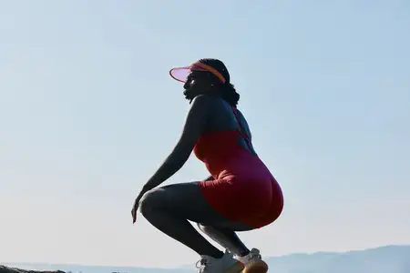 Подтянутая женщина в красном спортивном костюме приседает на каменистой поверхности у моря на фоне ясного голубого неба и холмов вдалеке