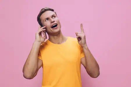 Весёлая женщина в ярко-жёлтой футболке прижимает мобильный телефон к уху, смотрит вверх и жестикулирует пальцем, розовый фон