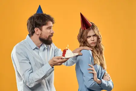 Мужчина и женщина в колпаках с тортом на жёлтом фоне, крупный план, празднование дня рождения