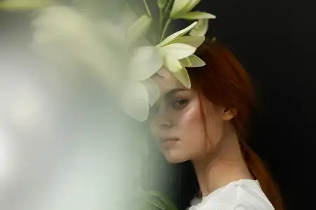 На этом портрете изображена молодая женщина с рыжими волосами, которую тонко обрамляют нежные белые лилии, демонстрируя гармоничное сочетание природы и элегантности в умиротворяющей обстановке