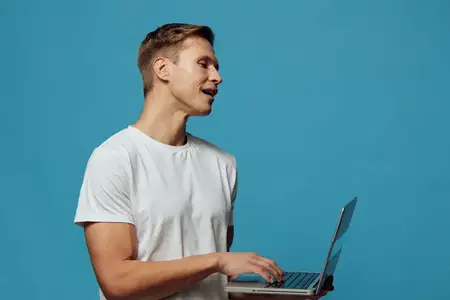 Счастливый мужчина с короткой стрижкой в повседневной белой футболке работает за ноутбуком на синем фоне