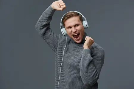 Взволнованный молодой мужчина в сером свитере и белых наушниках танцует и наслаждается музыкой с закрытыми глазами на темном фоне