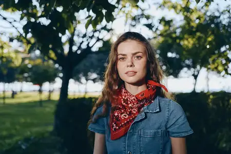 Женщина, ведущая активный образ жизни, одетая в современную джинсовую уличную одежду в стиле бохо-вестерн с красной банданой, стоит на открытом воздухе при естественном освещении в окружении зелени