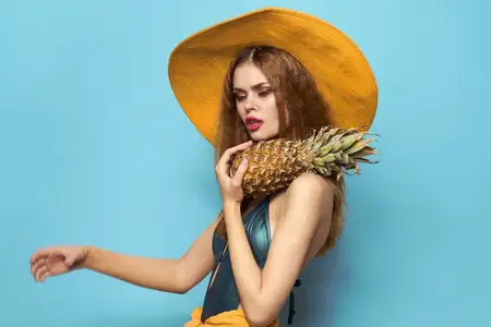 Женщина в пляжной шляпе держит ананас и купальник на фоне экзотических фруктов и голубого фона