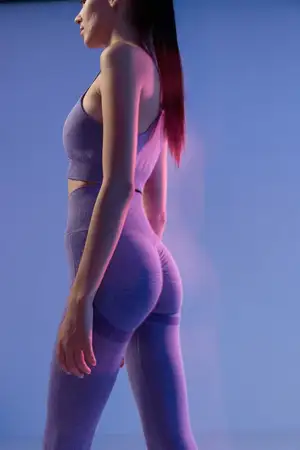 Женщина в фиолетовом бесшовном топе и леггинсах стоит в профиль в расслабленной позе на фоне плавного градиента синего и фиолетового цветов