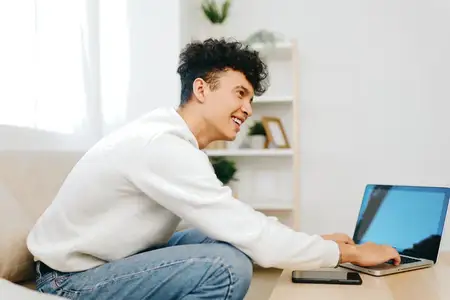 Улыбающийся молодой человек работает за ноутбуком, сидя на диване в хорошо освещённой комнате, демонстрируя непринуждённое и комфортное рабочее место с акцентом на продуктивность и современный…