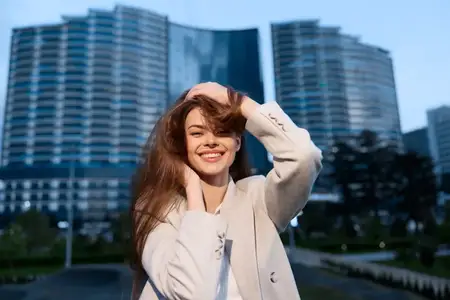 Радостная женщина в бежевом пальто уверенно улыбается на открытом воздухе рядом с современными небоскребами, передавая весенний сезон и городской образ жизни, деловая и модная тема