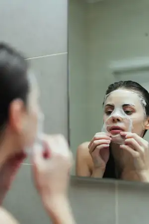 Сосредоточенная женщина выполняет процедуру ухода за кожей с маской для лица в хорошо освещённой ванной комнате, что подчёркивает заботу о себе и ритуалы красоты в домашних условиях