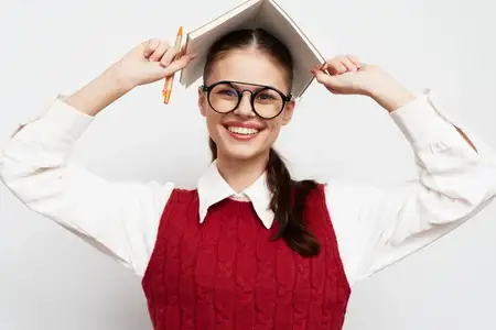 Радостная молодая женщина в очках и красном свитере игриво балансирует книгой на голове, её яркая улыбка создаёт живую атмосферу