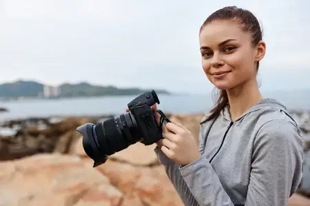 Счастливая молодая женщина в спортивной одежде держит камеру на пляже, воплощая радость фотографии и исследований на открытом воздухе, что идеально подходит для тем путешествий или образа жизни