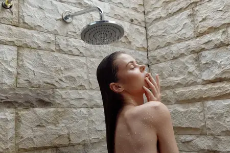 Молодая женщина стоит под уличным душем, капли воды на ее обнаженных плечах, глаза закрыты, безмятежное выражение лица, фон из натурального камня при дневном свете