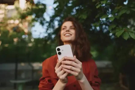 Улыбающаяся женщина стоит под деревьями, держа смартфон, одетая в повседневную красную куртку, естественный свет подчеркивает расслабленную повседневную сцену образа жизни