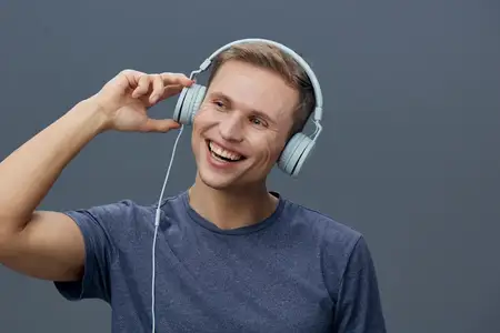 Счастливый молодой человек наслаждается музыкой в наушниках, в повседневной синей футболке, позитивное настроение, серый студийный фон, крупный план портрета