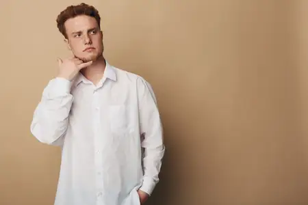 Задумчивая мужская модель в белой рубашке стоит, положив руку на подбородок, и смотрит в сторону на бежевом фоне