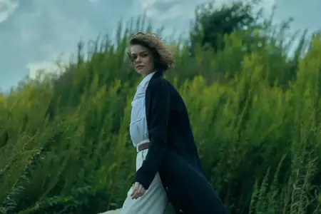 Модное движение и ветер в поле: женщина шагает вперед, пальто развевается, современный стиль среди зеленой травы и открытого неба
