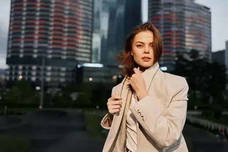 Стильная женщина в бежевом пальто уверенно стоит на открытом воздухе на фоне современного городского пейзажа, при естественном освещении и осенней атмосфере, выражая спокойствие и решимость