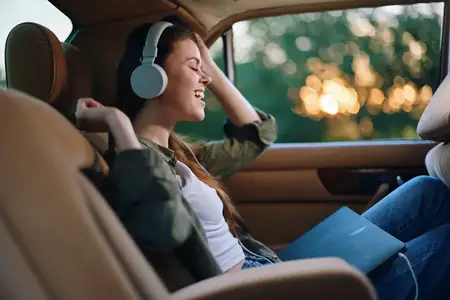 Радостная молодая женщина слушает музыку в автомобиле, надев стильные наушники и повседневную одежду