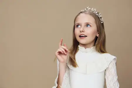 Очаровательная маленькая девочка в тиаре и красивом белом платье, излучающая радость и любопытство, что идеально подходит для тем детских воспоминаний, праздников и семьи, на фоне бежевого фона