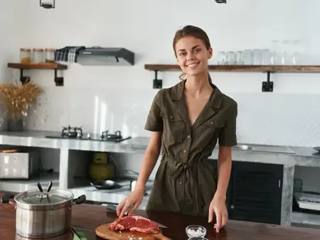 Женщина с улыбкой на кухне готовит обед из мяса, овощей и специй на деревянном столе на фоне стильной кухни