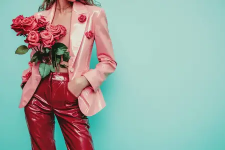 Модная модель в розовом блейзере и блестящих красных брюках, держащая букет роз, излучающая радость и летнее настроение на фоне яркой бирюзовой стены