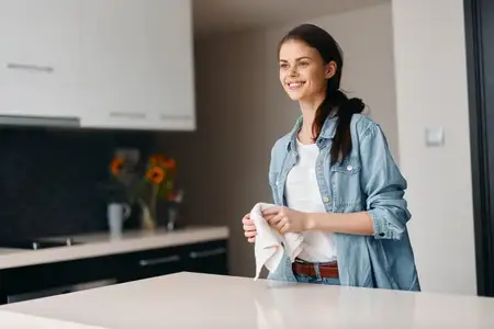 Счастливая молодая женщина наслаждается домашним уютом на современной кухне: домохозяйка европеоидной внешности улыбается, убираясь с моющим средством